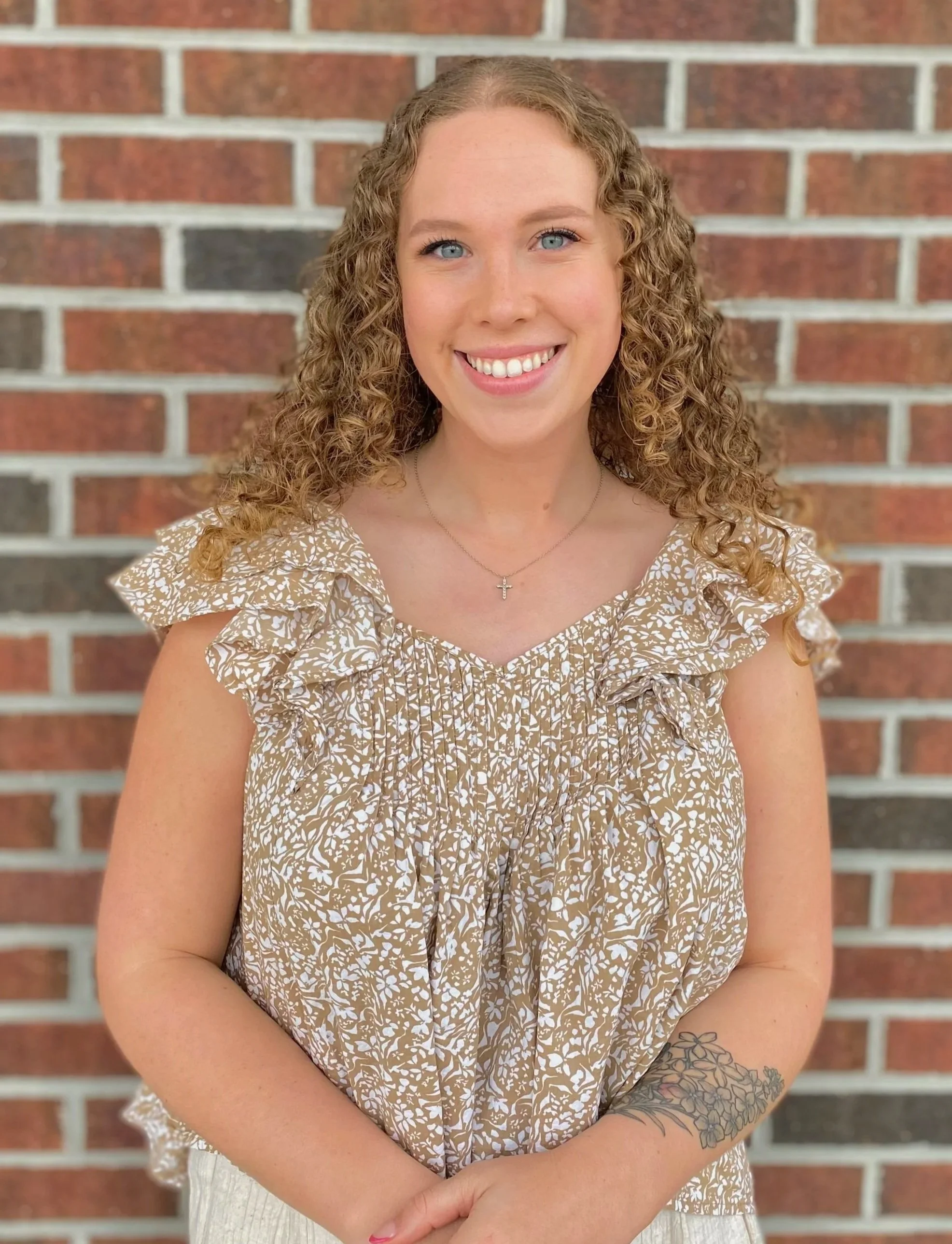 Woman with curly hair smiling at the beach, wearing a beige tank top and patterned pants, with a visible tattoo on her left arm. A beach house and a blue sky are in the background.