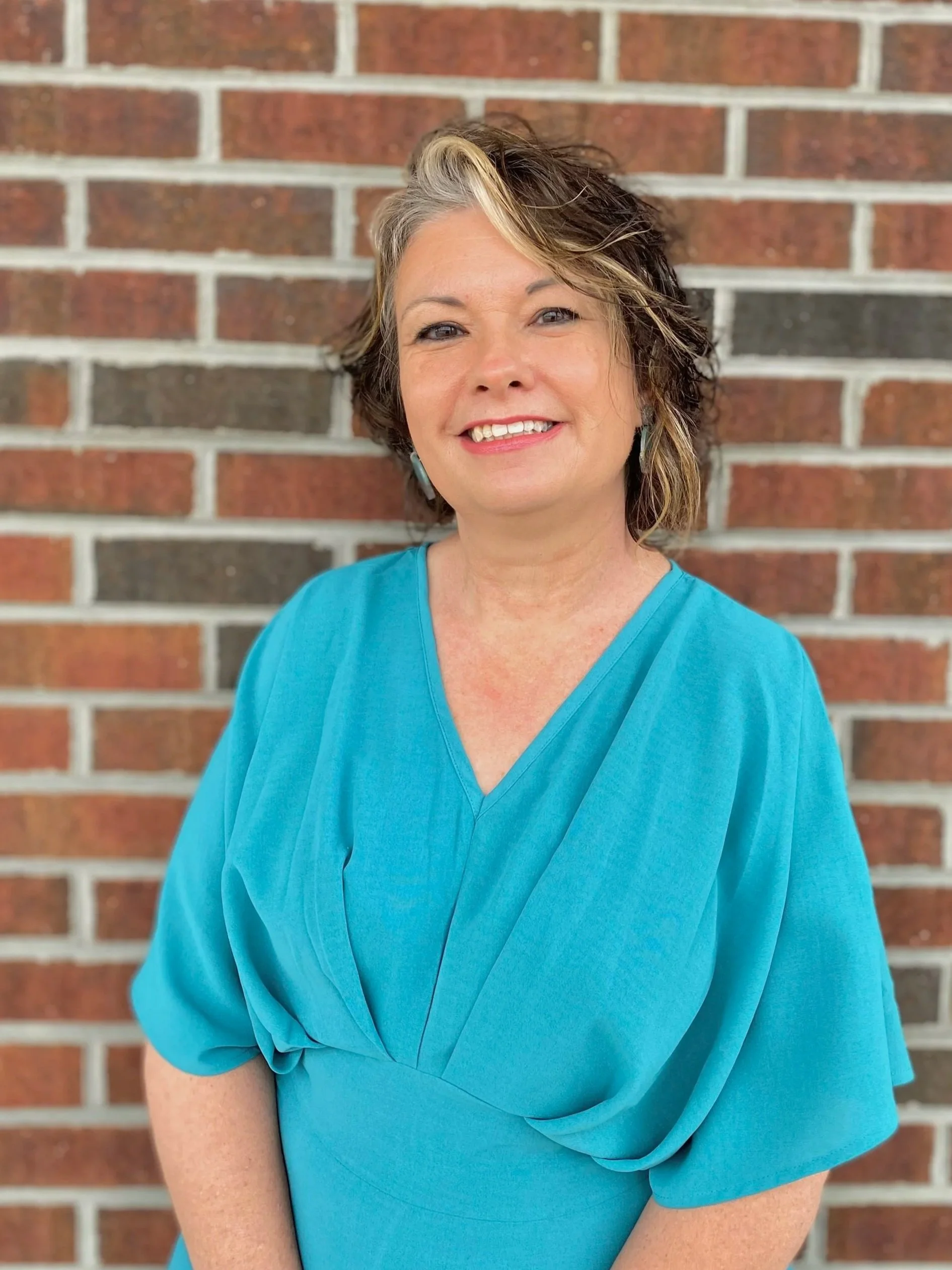Woman wearing a green dress and gold hoop earrings, smiling in front of a white shiplap wall.