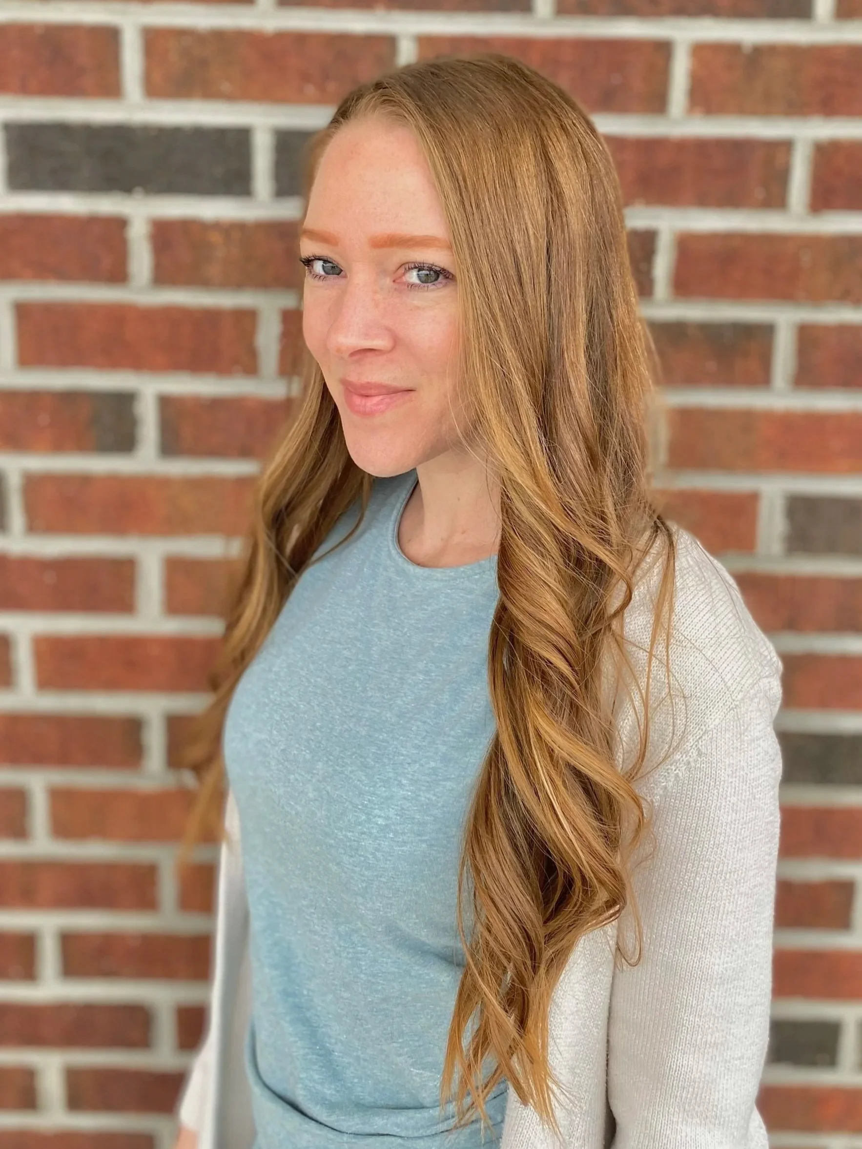 Woman with long red hair wearing a white sleeveless top, turquoise pants, layered necklace, and a smartwatch, standing in front of a light-colored shiplap wall.