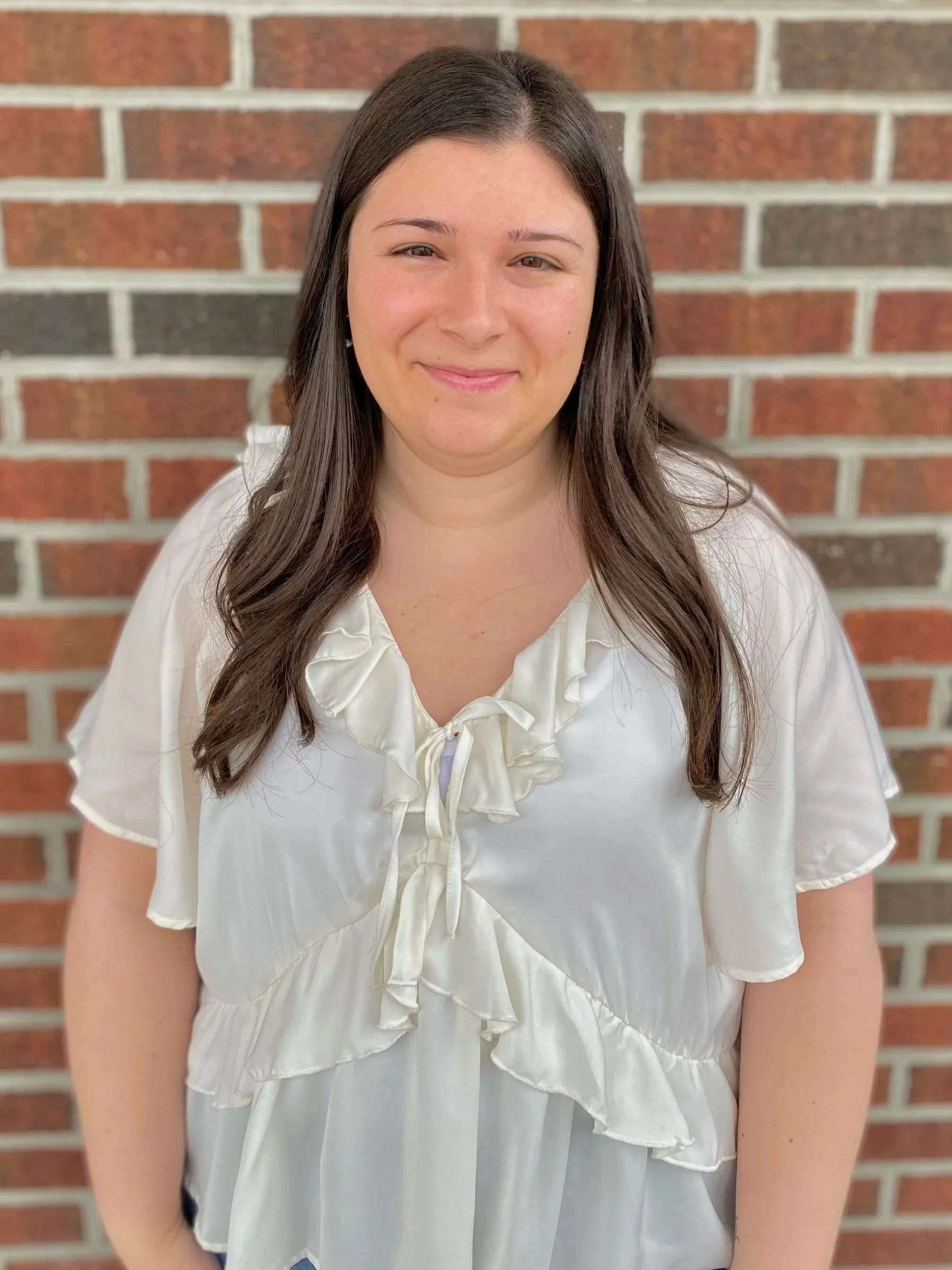 Woman in green top and white pants standing against a shingle wall, smiling.