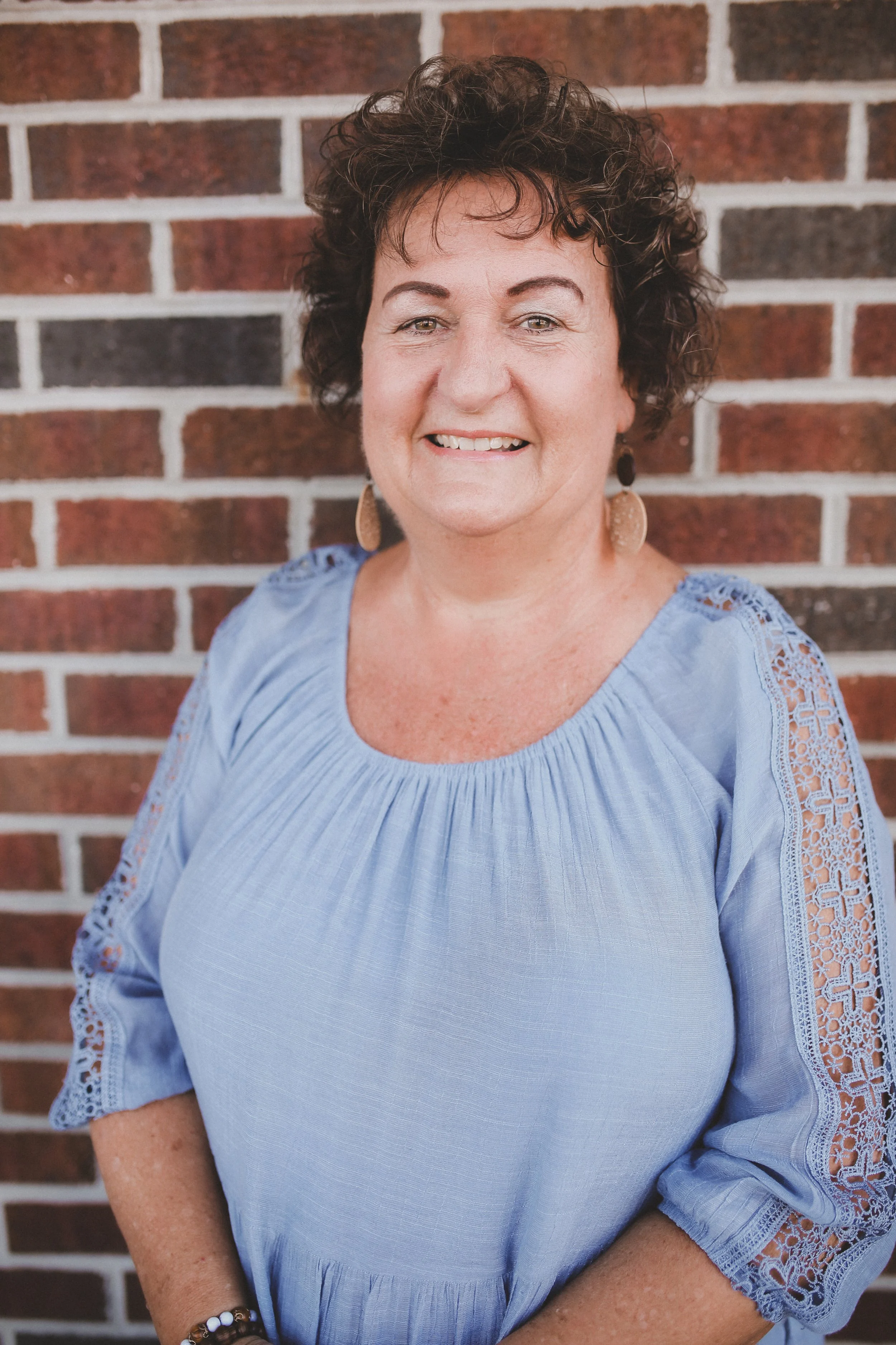 Person wearing a light blue blouse with a starfish necklace, standing against a wood-paneled wall.