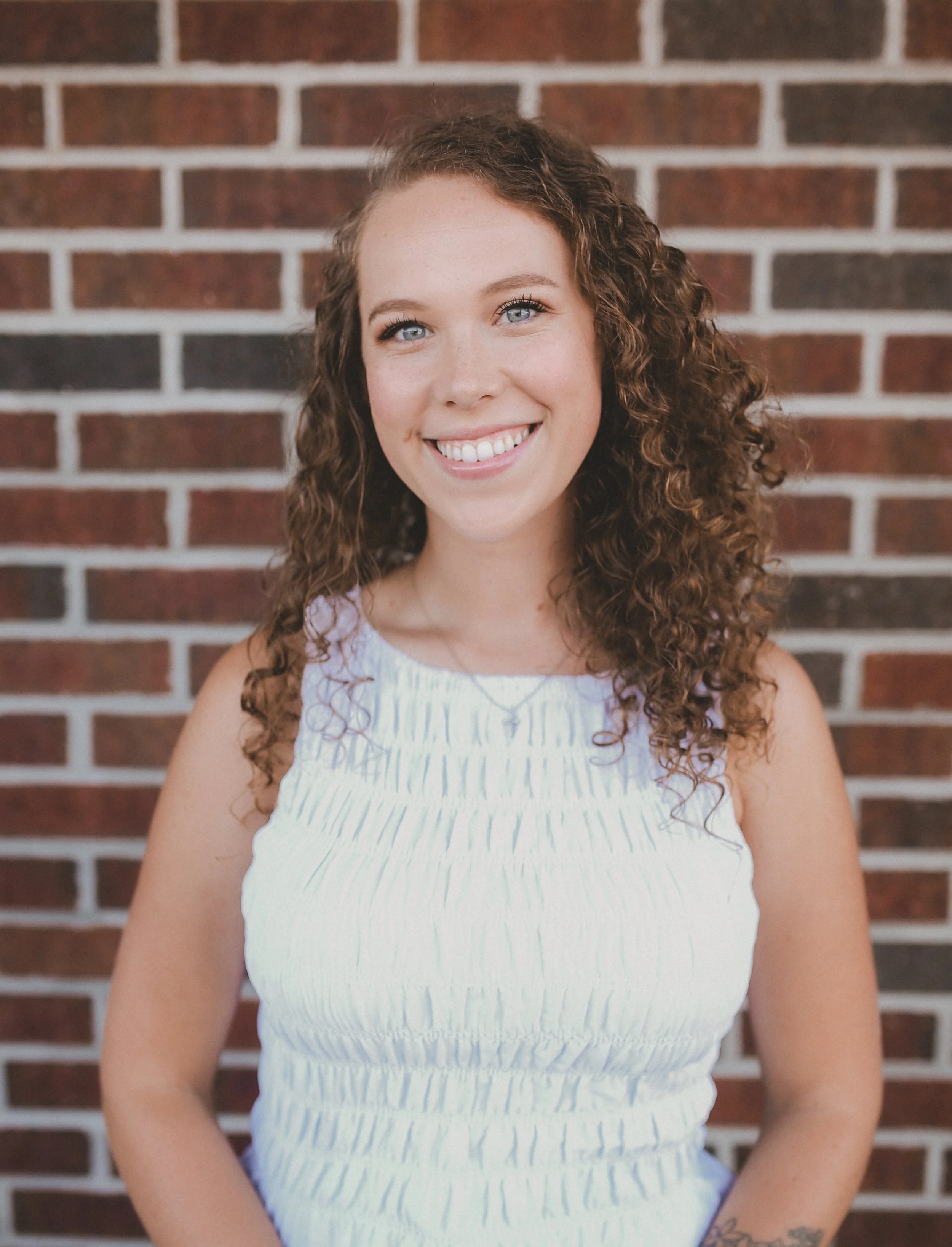 Woman with curly hair smiling at the beach, wearing a beige tank top and patterned pants, with a visible tattoo on her left arm. A beach house and a blue sky are in the background.