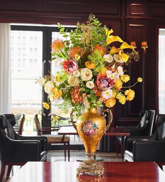 A large arranged bouquet of colorful flowers in a decorative gold vase on a wooden table in a well-lit room with dark wood-paneled walls and black leather chairs.