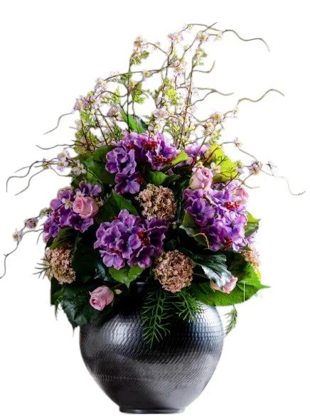 A floral arrangement with purple, pink, and beige flowers in a black textured vase, accented with curly branches and green foliage.
