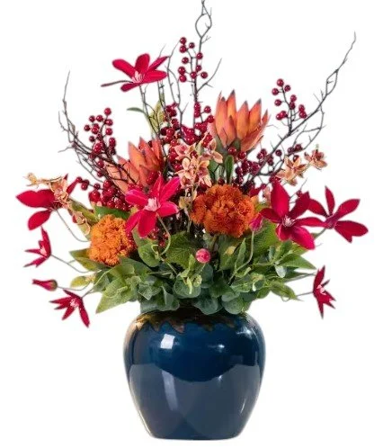 Colorful floral arrangement in a dark blue vase, featuring red, orange, and pink flowers with green leaves and decorative branches.