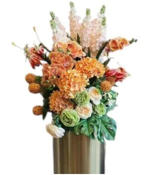 A large floral arrangement featuring orange, peach, white, and green flowers in a tall, cylindrical gold vase.