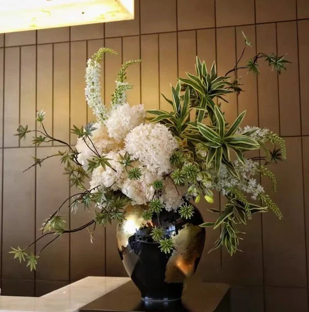 A floral arrangement in a black glossy vase on a table, featuring white hydrangeas, green foliage with variegated leaves, and white flowers, set against a wooden-paneled wall.