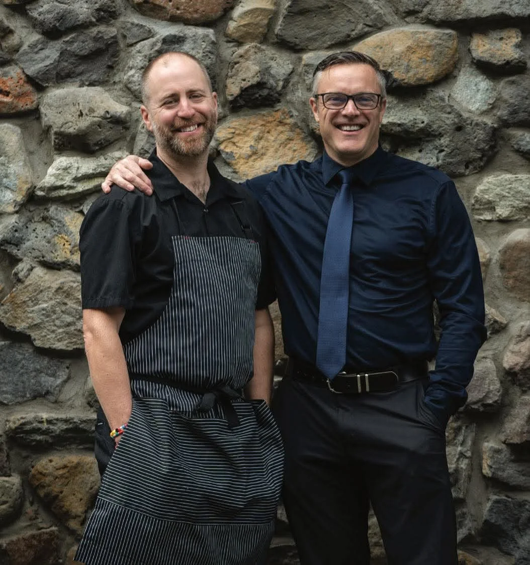 Two men standing in front of a stone wall, one wearing a striped apron and the other in a dark shirt and tie, smiling.