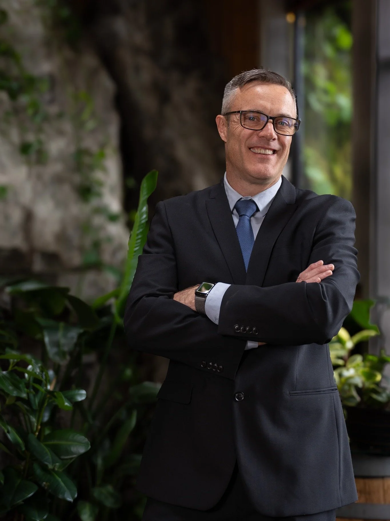 A smiling man in a black suit, blue tie, and glasses, standing with arms crossed in front of a window with greenery outside.