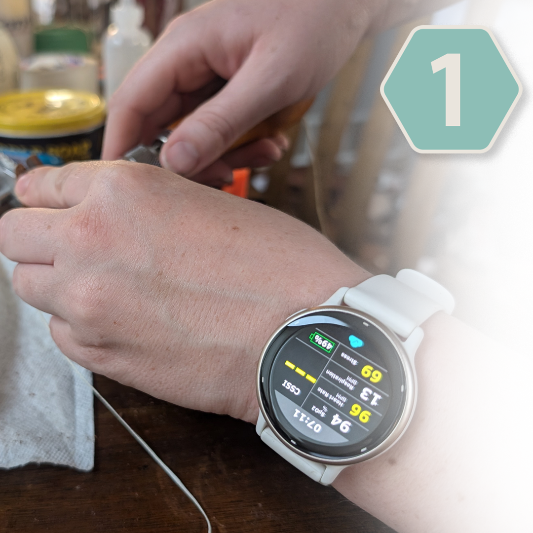 A person wearing a white smartwatch on their left wrist, showing health data on the screen, with their right hand holding an object, possibly a tool, over a wooden work surface. In the background, there are household items including a container with a yellow lid.