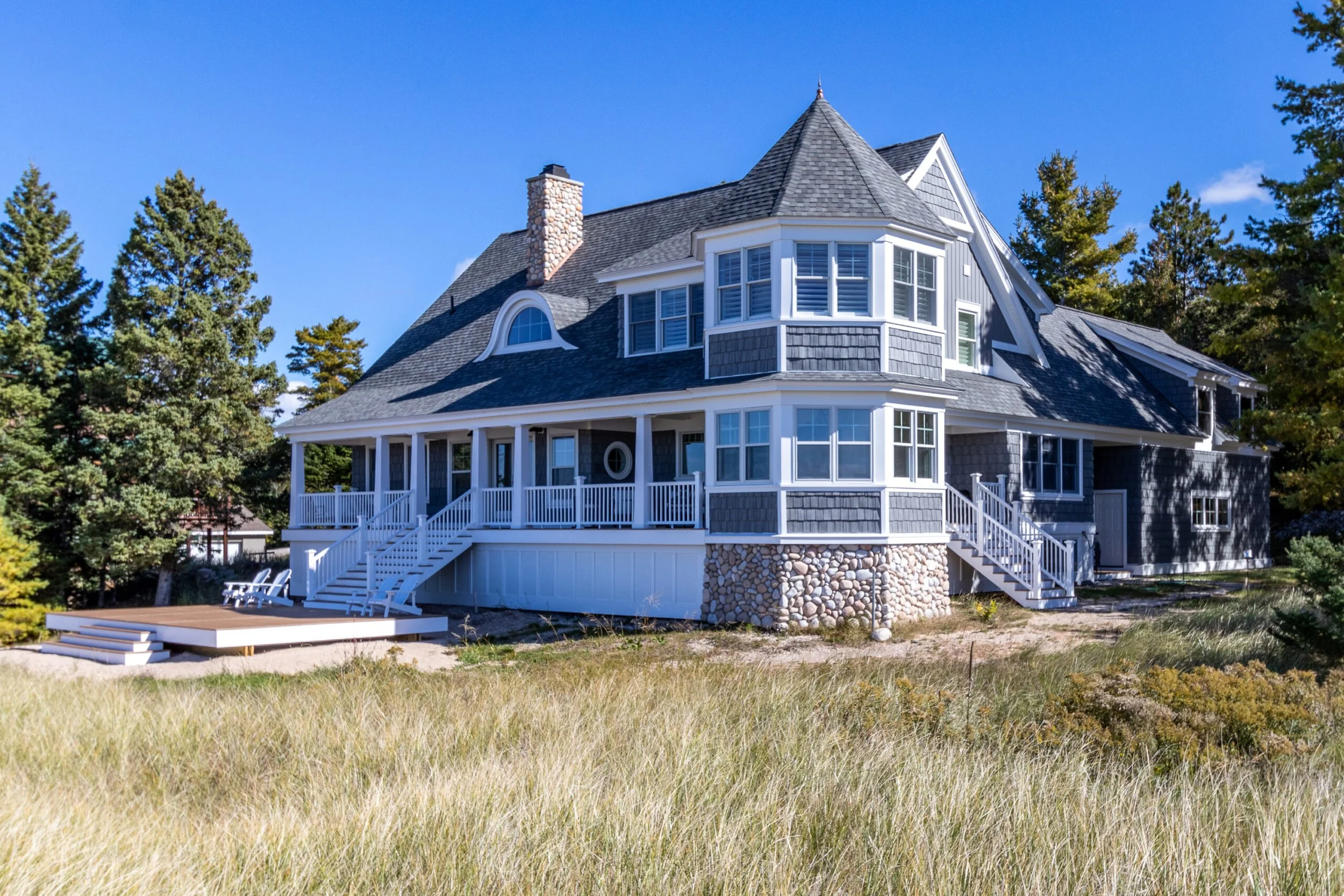 A large, multi-story house with a steep roof and multiple windows, surrounded by trees and grass, under a clear blue sky.