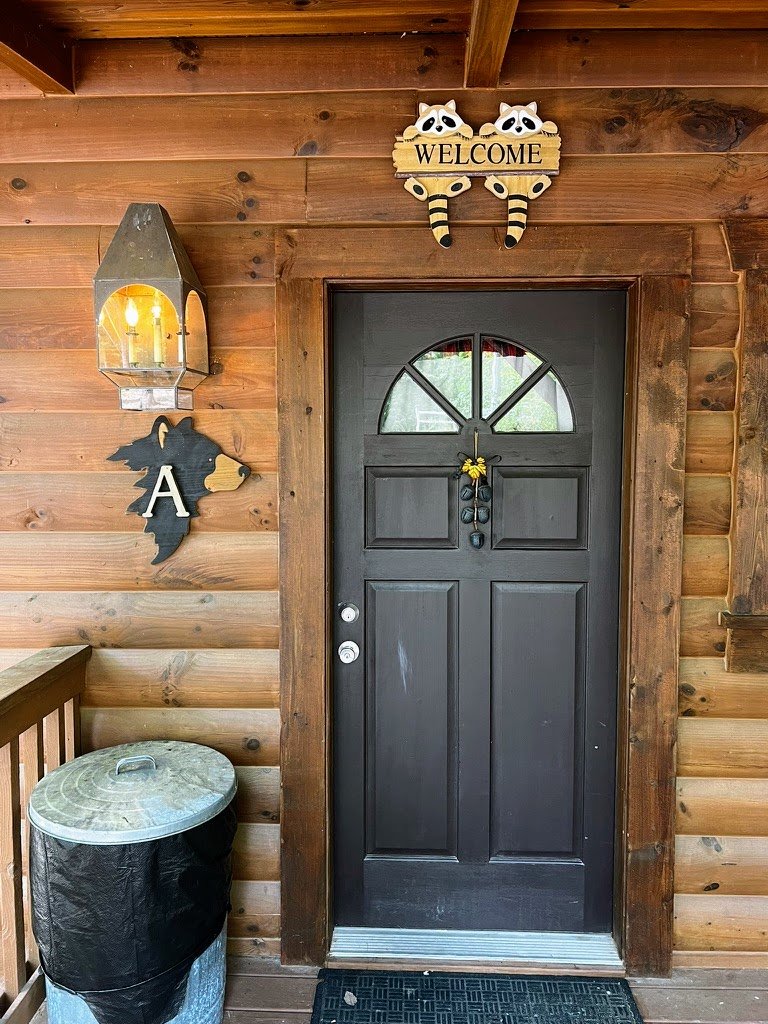 Front entrance of a wooden cabin with a black door, raccoon-themed "Welcome" sign above, and decorative items such as a lantern and bear silhouette. A metal trash can is on the left side.