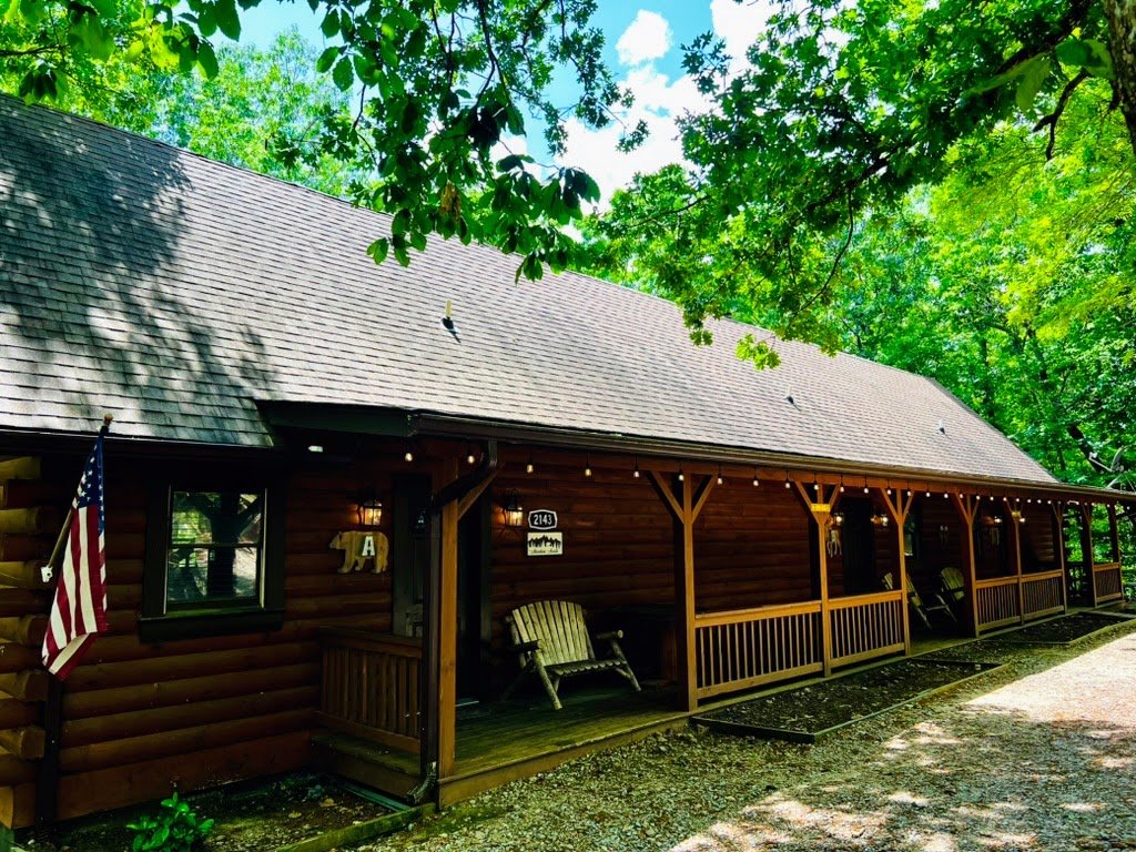 A rustic log cabin with a porch, surrounded by trees. An American flag is displayed near the entrance. Wooden chairs are placed on the porch, under a string of lights.
