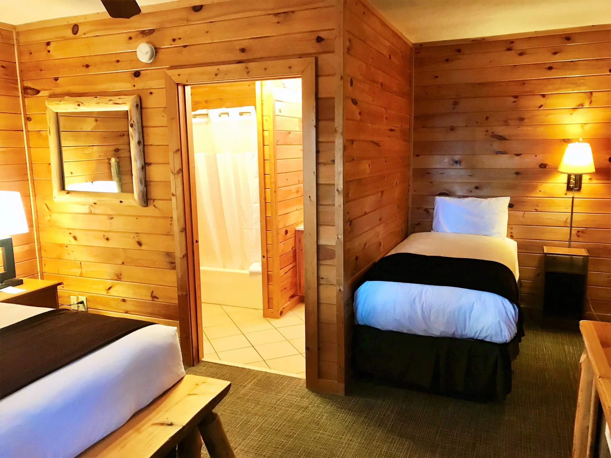 Rustic cabin-style hotel room with wood-paneled walls, featuring a twin bed, a larger bed partially visible, a nightstand with a lamp, a large mirror, and an open door to a tiled bathroom with a shower.