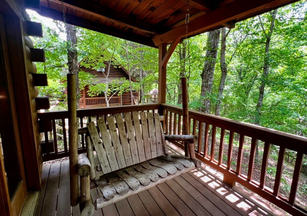Wooden porch swing on a cabin balcony overlooking trees and another cabin.