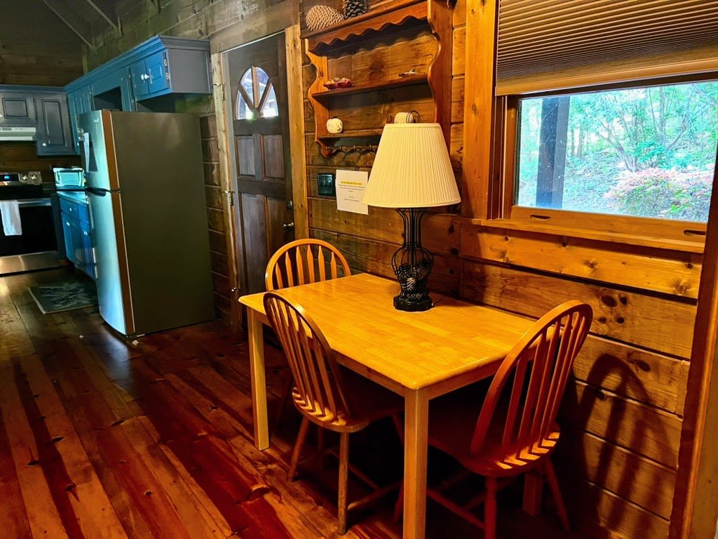 Rustic kitchen setting with wooden walls, dining table with two chairs, a lamp, and a window showing greenery outside. Kitchen features include blue cabinets and a stainless steel refrigerator.