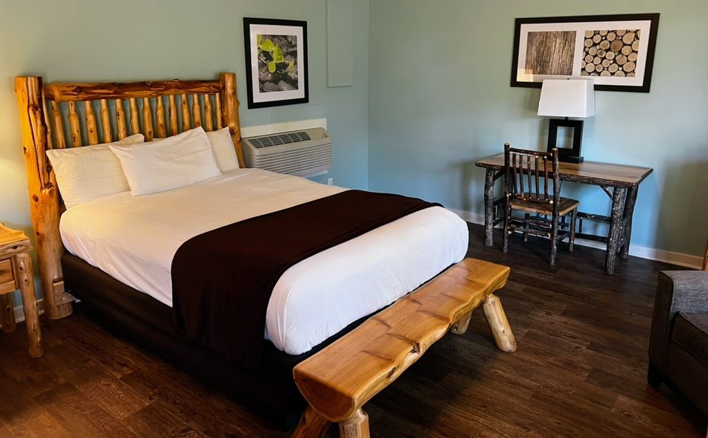 Rustic hotel room with a wooden bed frame, white bedding, brown throw blanket, a wooden bench at the foot of the bed, a wooden desk and chair, wall art, a lamp, and hardwood flooring.
