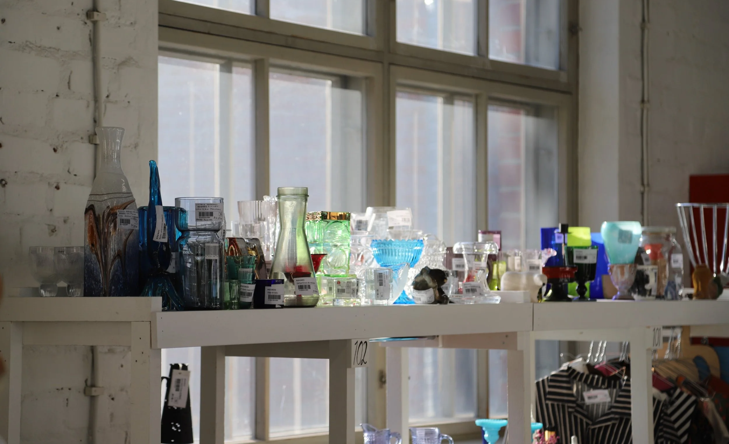 Värikkäitä vintage-lasiesineitä kauniisti esillä Round2 Thriftshop and Coffee -kirpputorin pöydällä Jyväskylässä, luonnonvalon osuessa teollisen tilan ikkunoista.