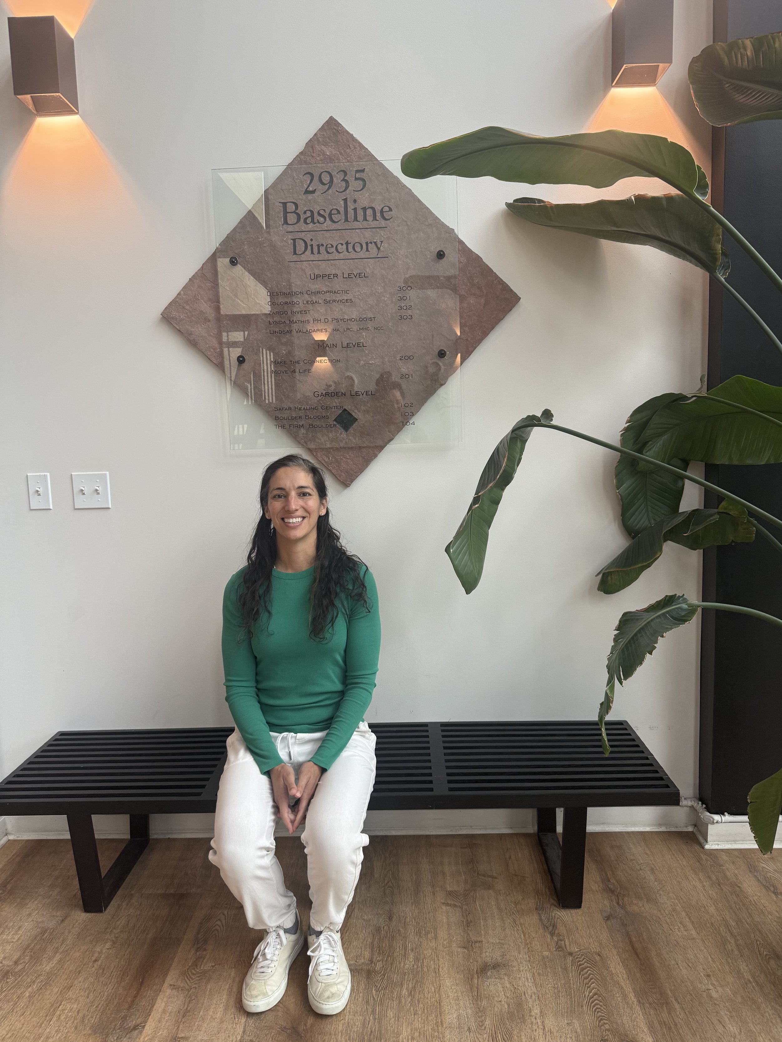Dr Leslie sitting on a bench in front of the directory signage in the lobby of the building for Destination Chiropractic in Boulder, Colorado.