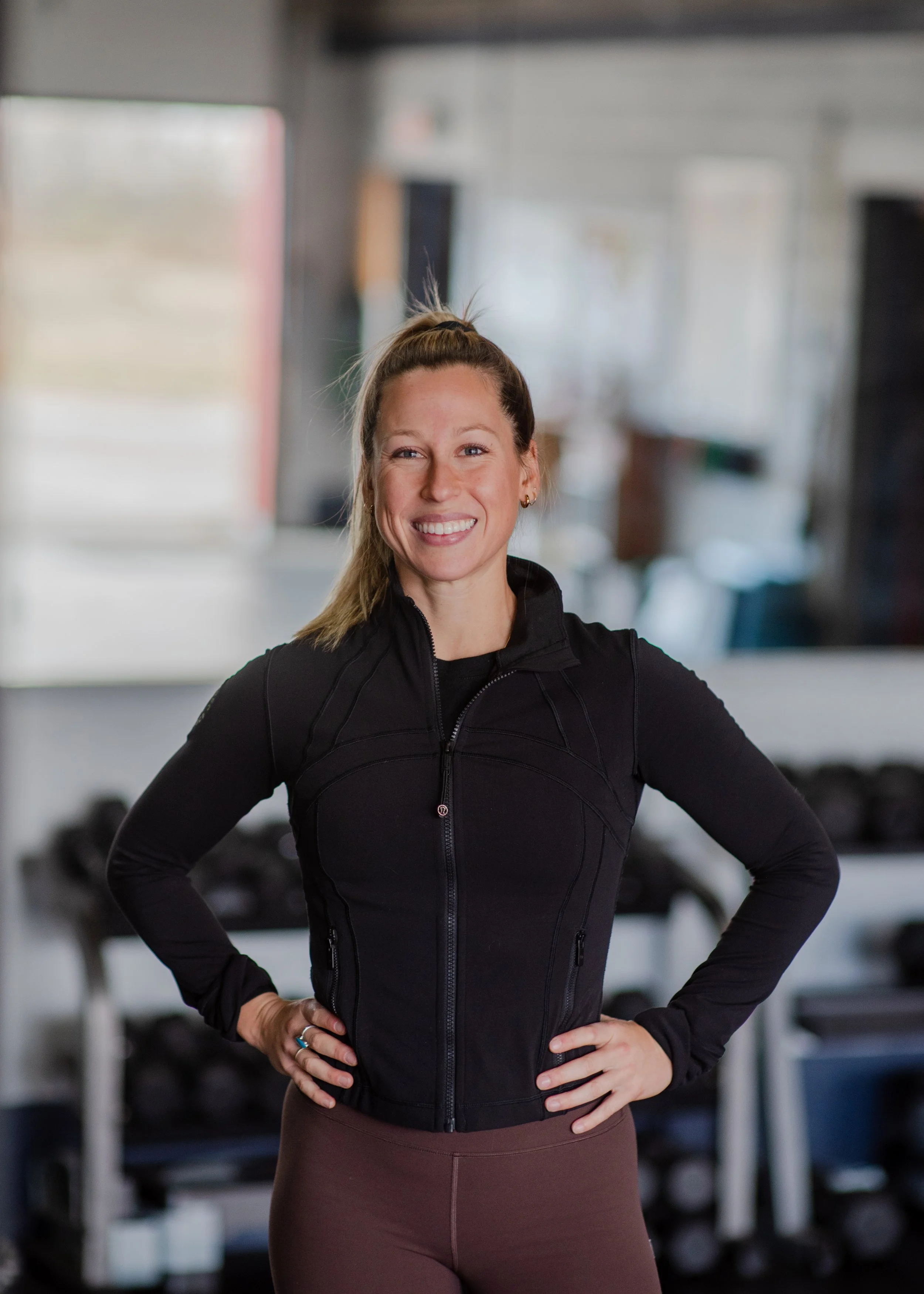 A woman smiling with hands on her hips, wearing a black athletic jacket and brown leggings, standing in a gym.