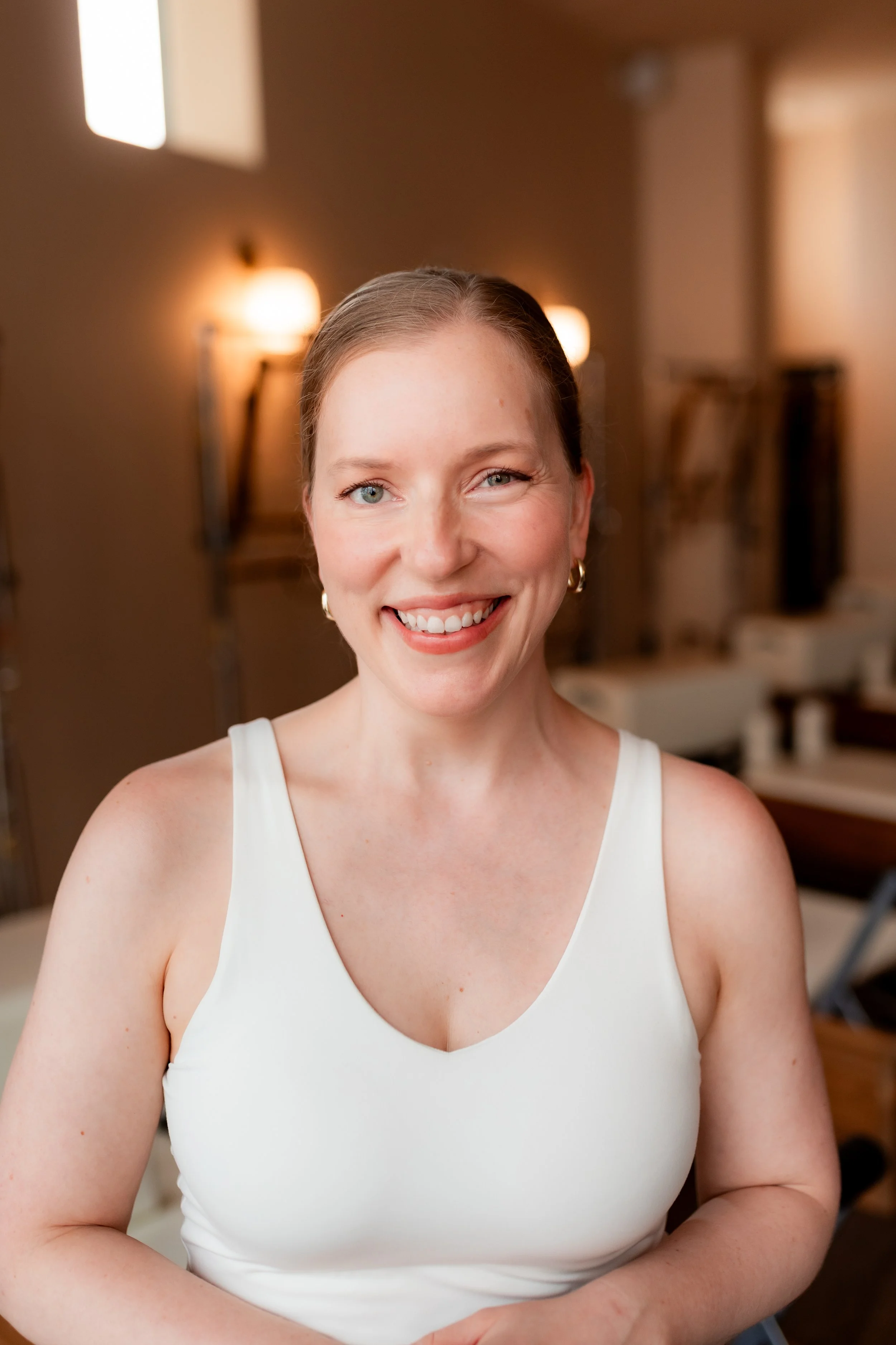 A woman with long light brown hair smiling in a room with a peach-colored wall and various electronic equipment and furniture in the background.