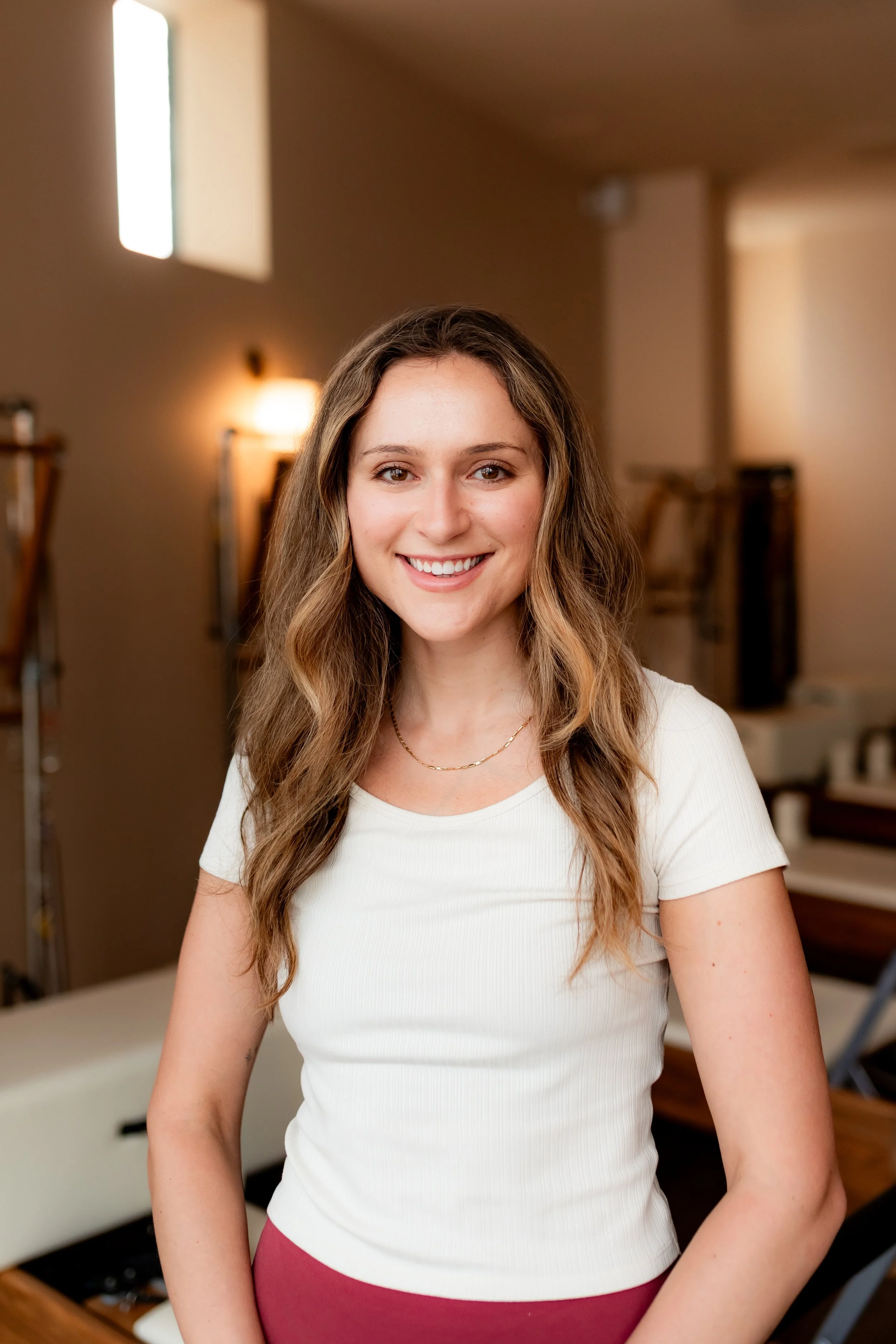 A young woman with long brown hair smiling in a room with beige walls and warm lighting. She is wearing a gray t-shirt and black pants, with a brown bracelet on her wrist.