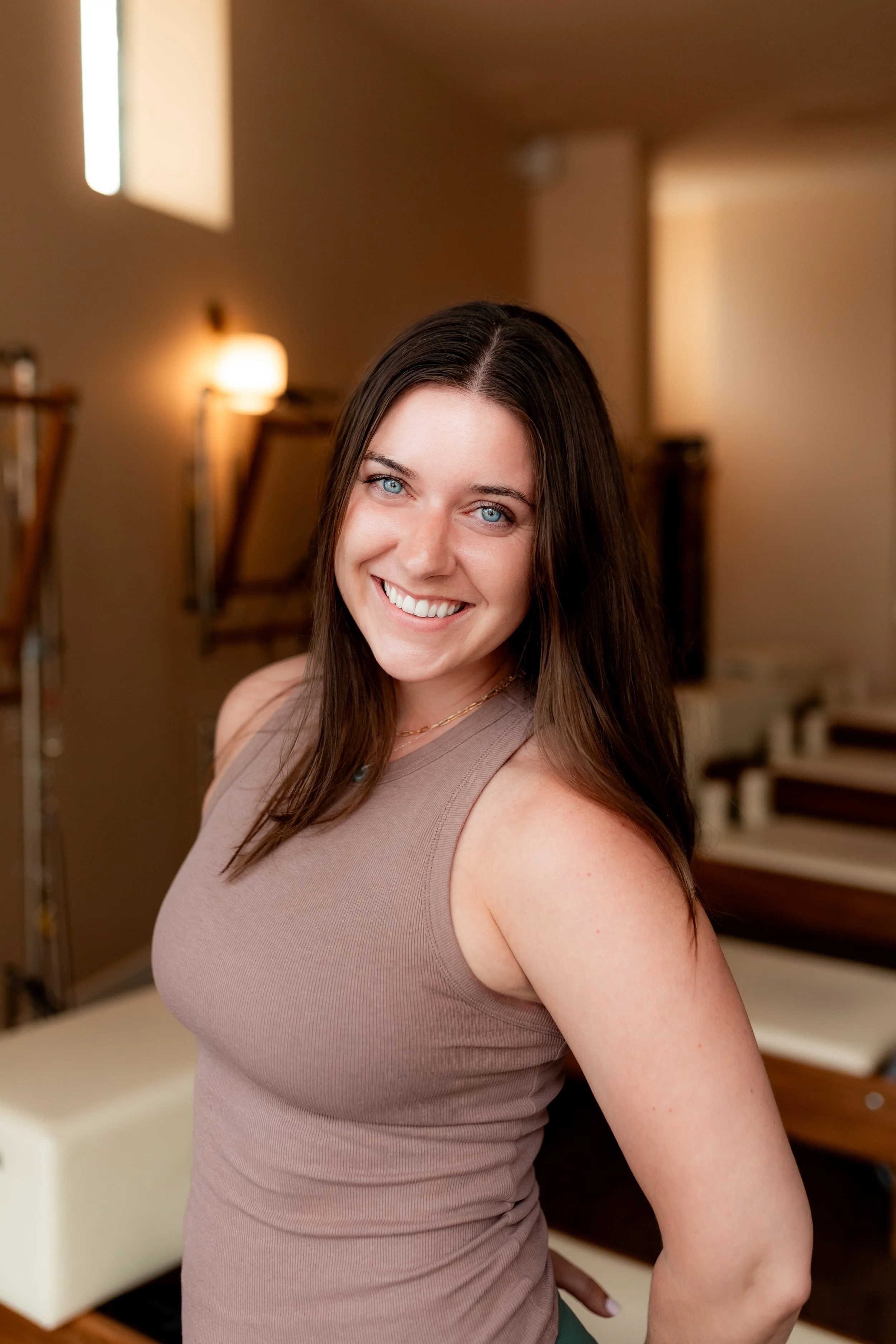 A young woman with long brown hair and blue eyes smiling in a living room, wearing a dark long sleeve top and teal pants.