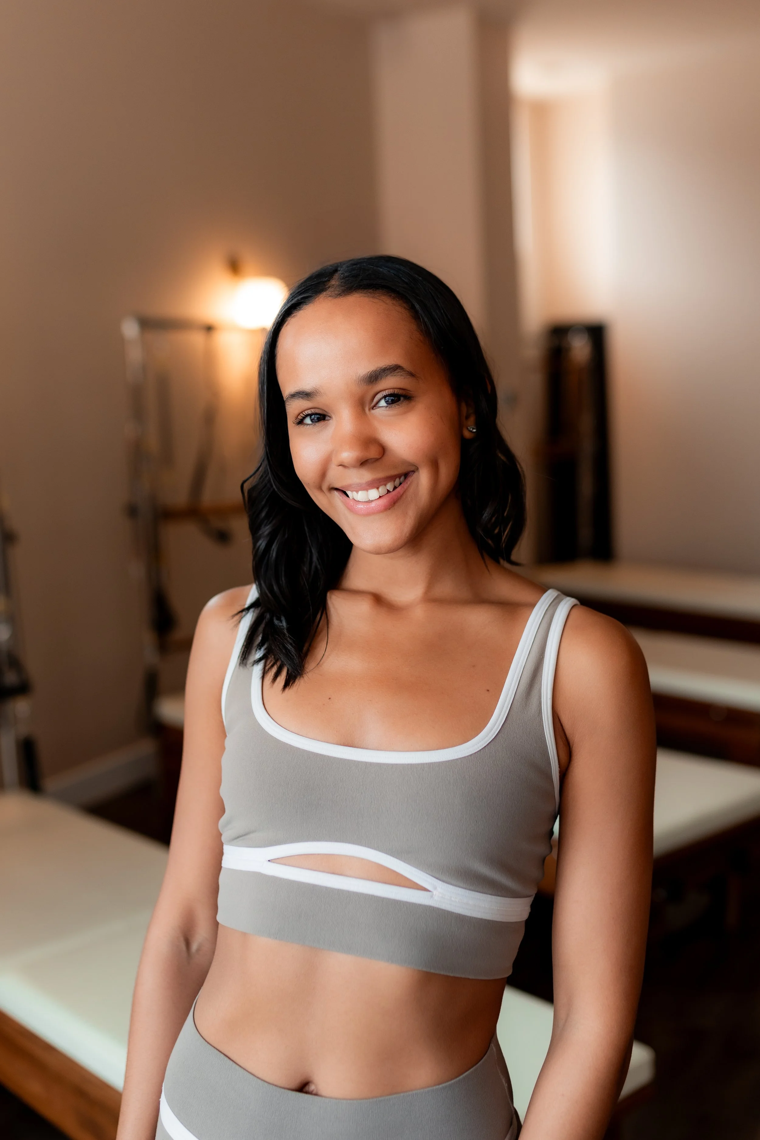 A smiling girl standing in a Pilates or fitness studio with equipment and soft lighting.