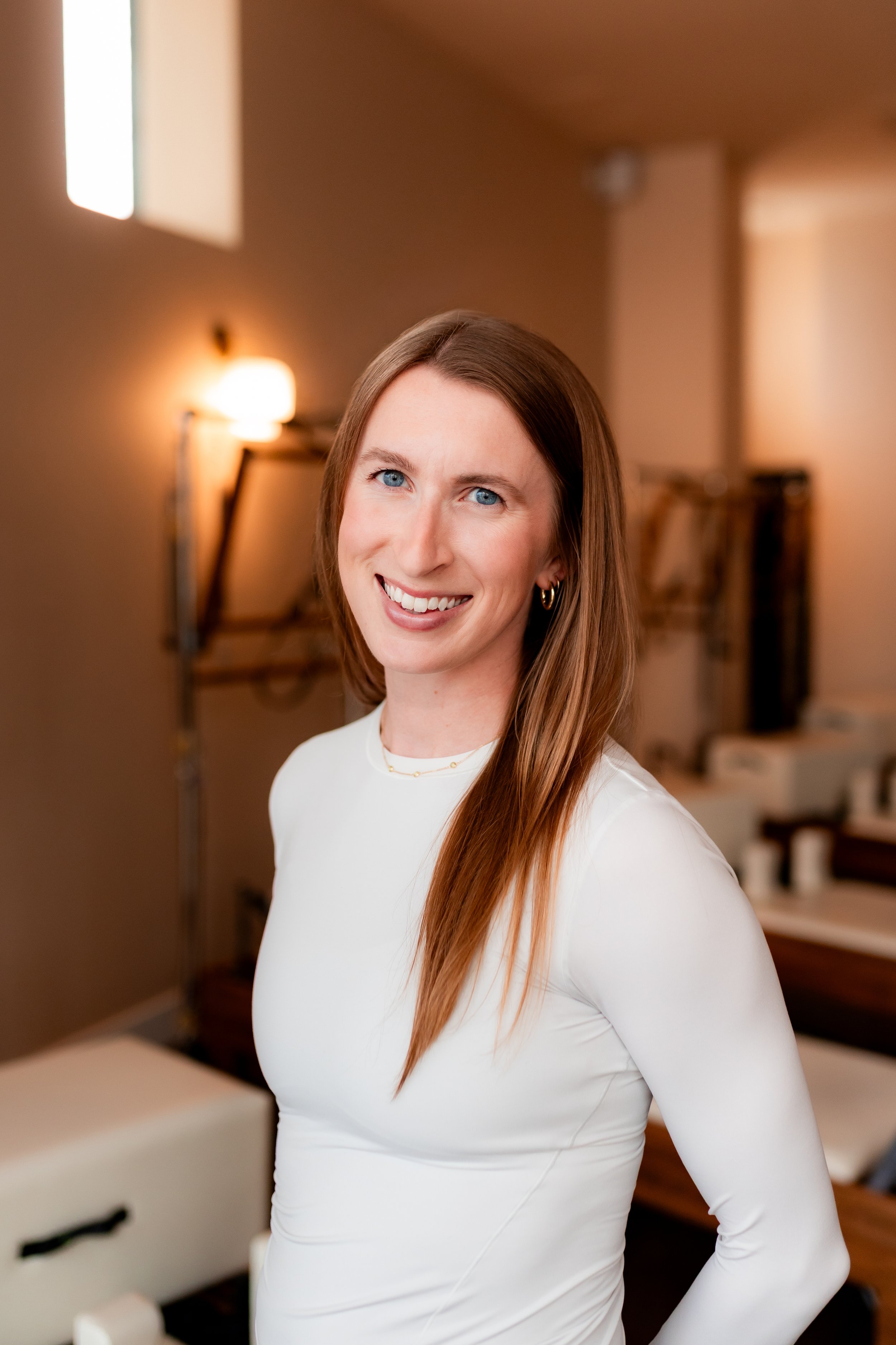 A woman with long brown hair and blue eyes smiling, standing in a gym or fitness studio, wearing a dark workout outfit.