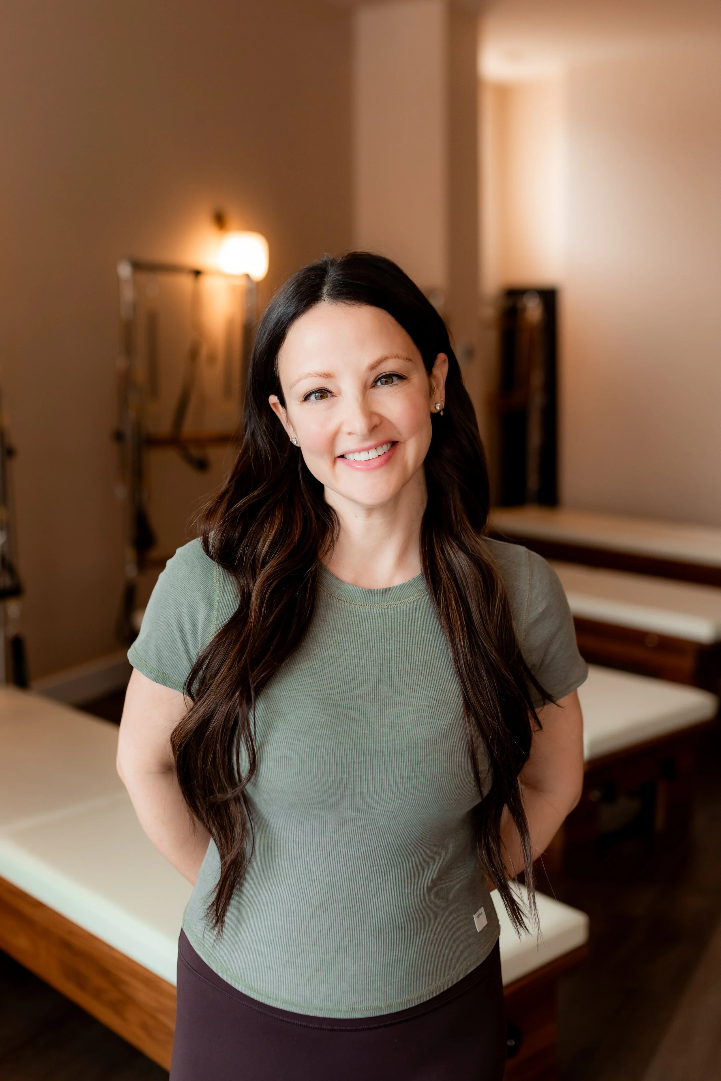 A woman with long dark hair and hoop earrings smiling indoors, wearing a purple sports bra with the letters 'SET' on it, in a room with warm lighting and art on the walls.