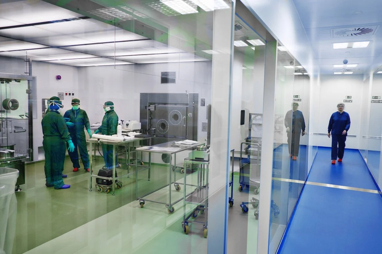 Hospital operating room with medical staff in green scrubs and masks, viewed through glass walls, and a corridor with two medical professionals walking.