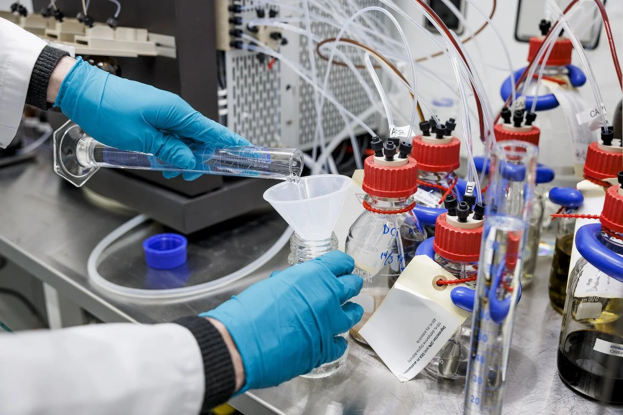 A scientist wearing blue gloves is pouring a liquid into a glass funnel in a laboratory with various laboratory bottles and equipment.