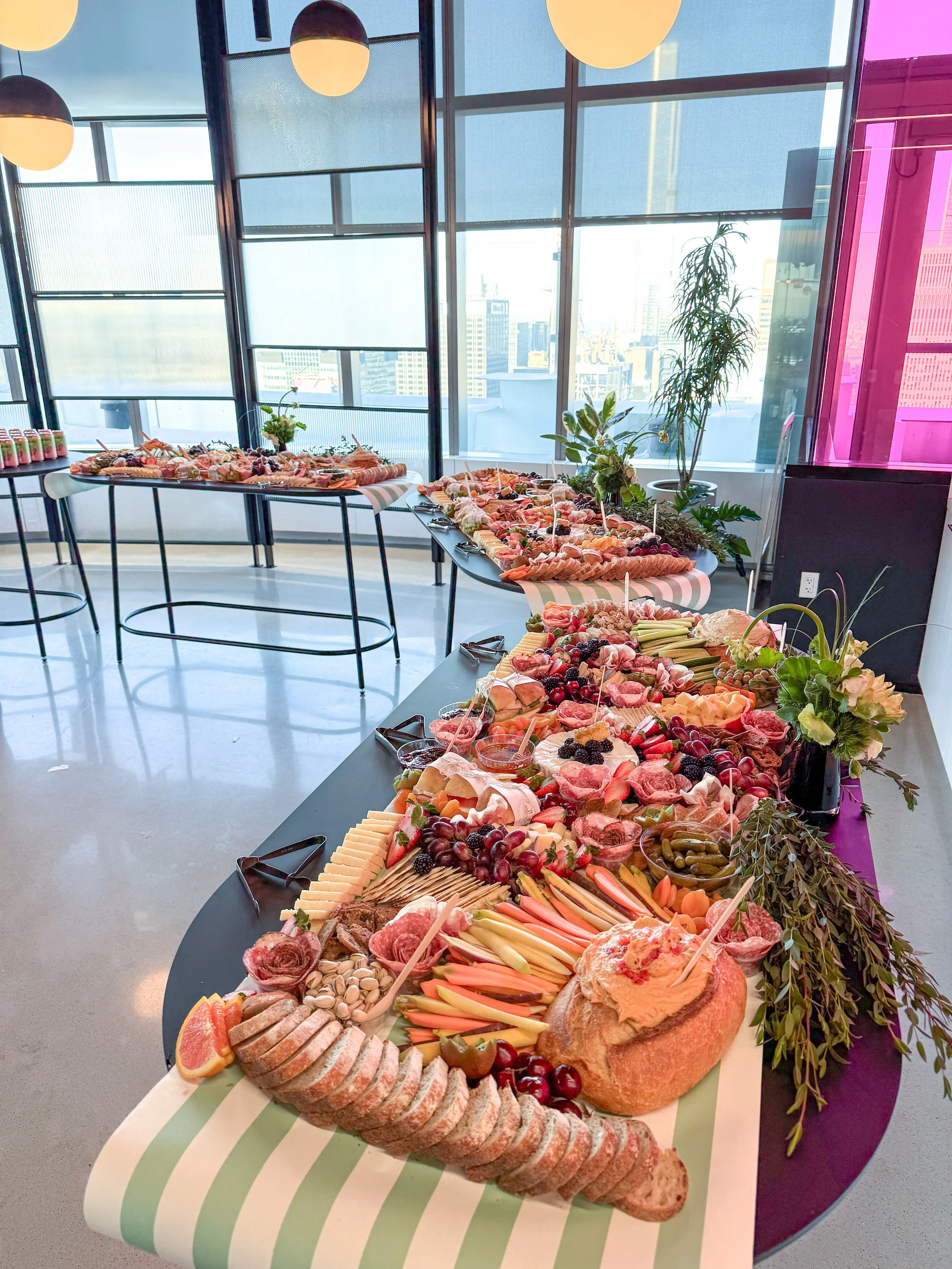Buffet table with various meats, cheeses, fruits, vegetables, and bread, set in a bright room with large windows and modern decor.