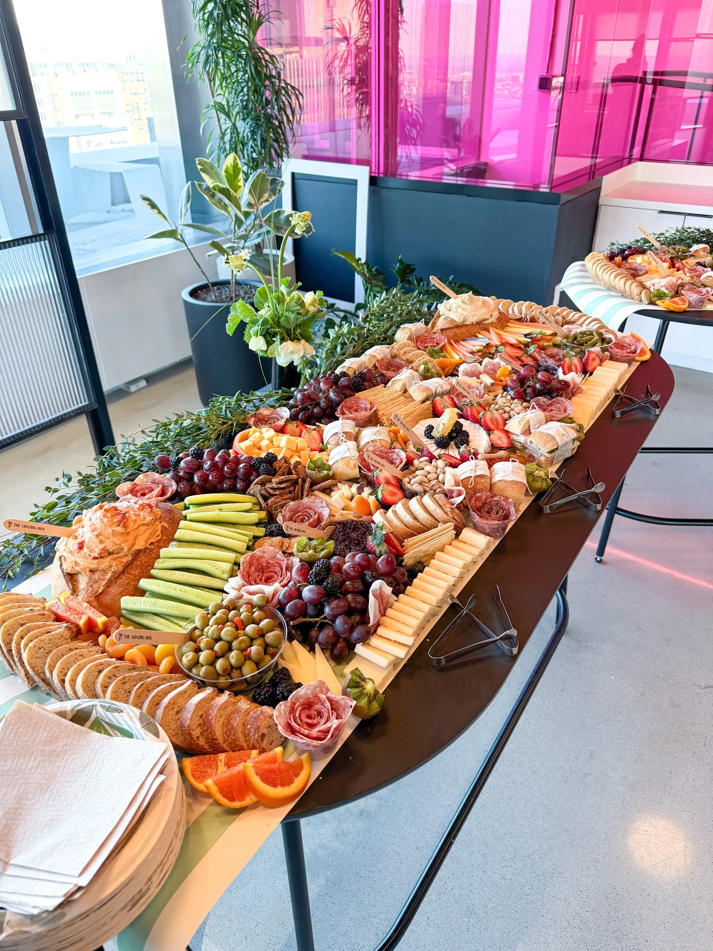 A large tray of assorted fruits, cheeses, and snacks on a black table in a bright modern room with plants, a view of the city, and pink wall panels.