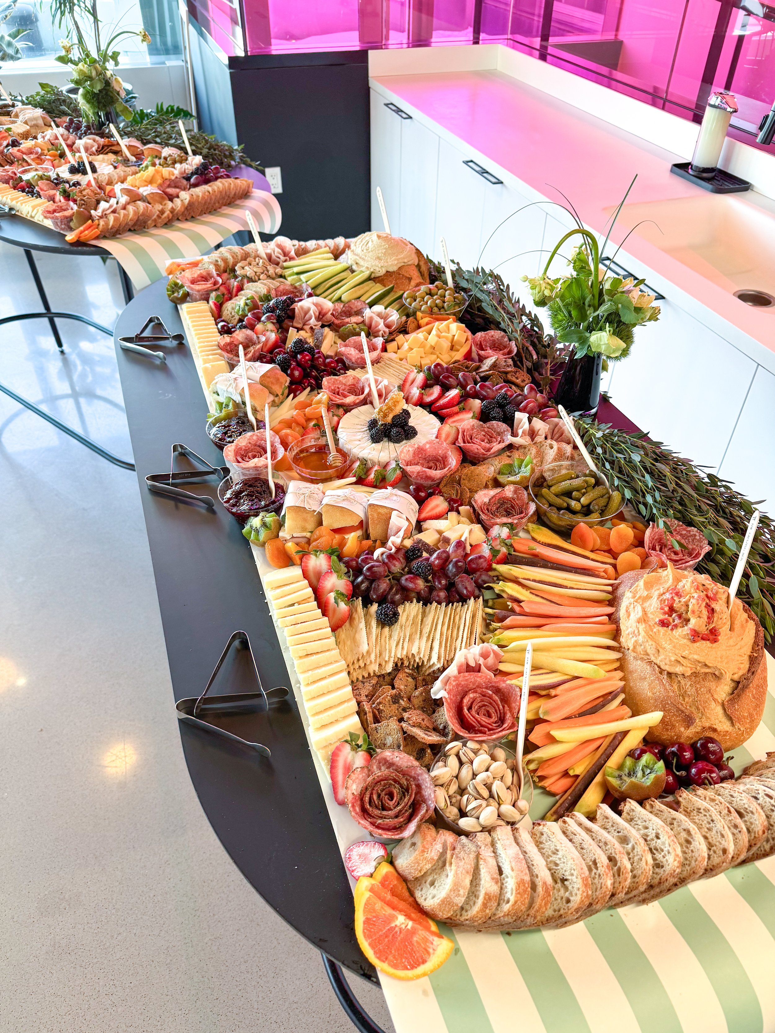 Large charcuterie and fruit platter with cheeses, grapes, strawberries, sliced oranges, and assorted snacks, on a black table with tongs.