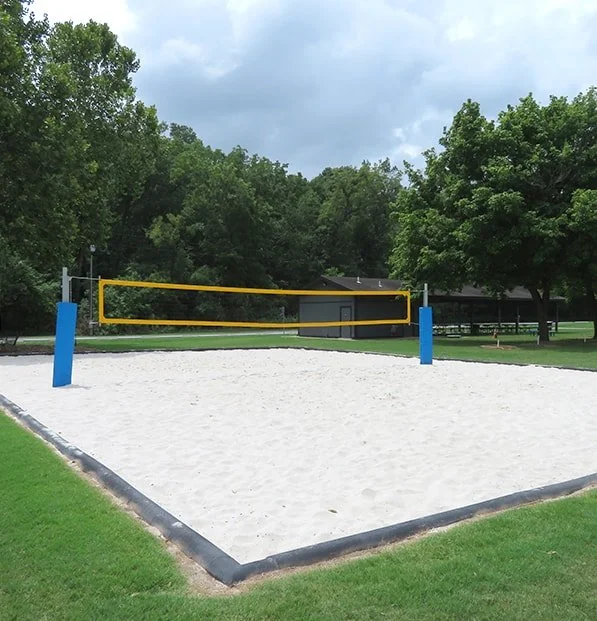 Outdoor sand volleyball court with a yellow net and blue poles surrounded by grass and trees under a cloudy sky.