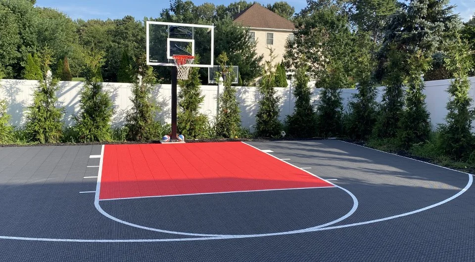 Outdoor basketball half-court with red and gray flooring, surrounded by a white fence and greenery.