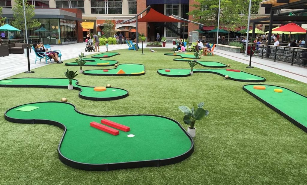 Outdoor mini-golf course with artificial grass, multiple holes, and people enjoying the activity in a lively urban setting.