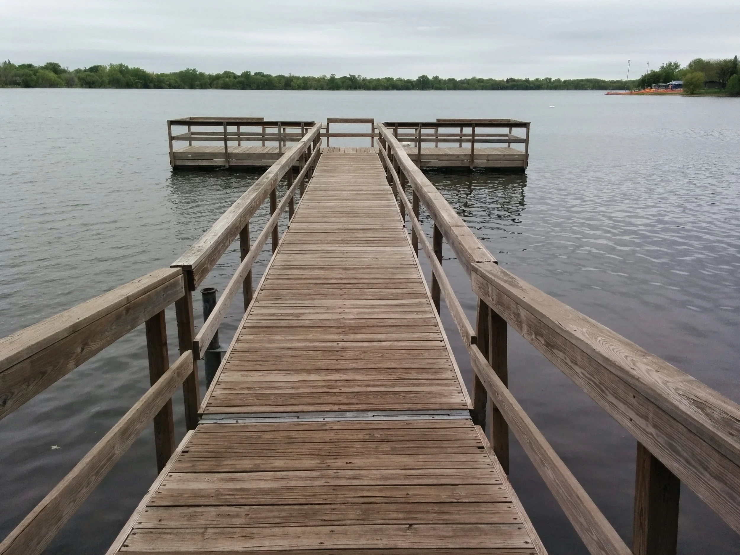 Wooden dock with railing extending over a calm lake, surrounded by trees under a cloudy sky.