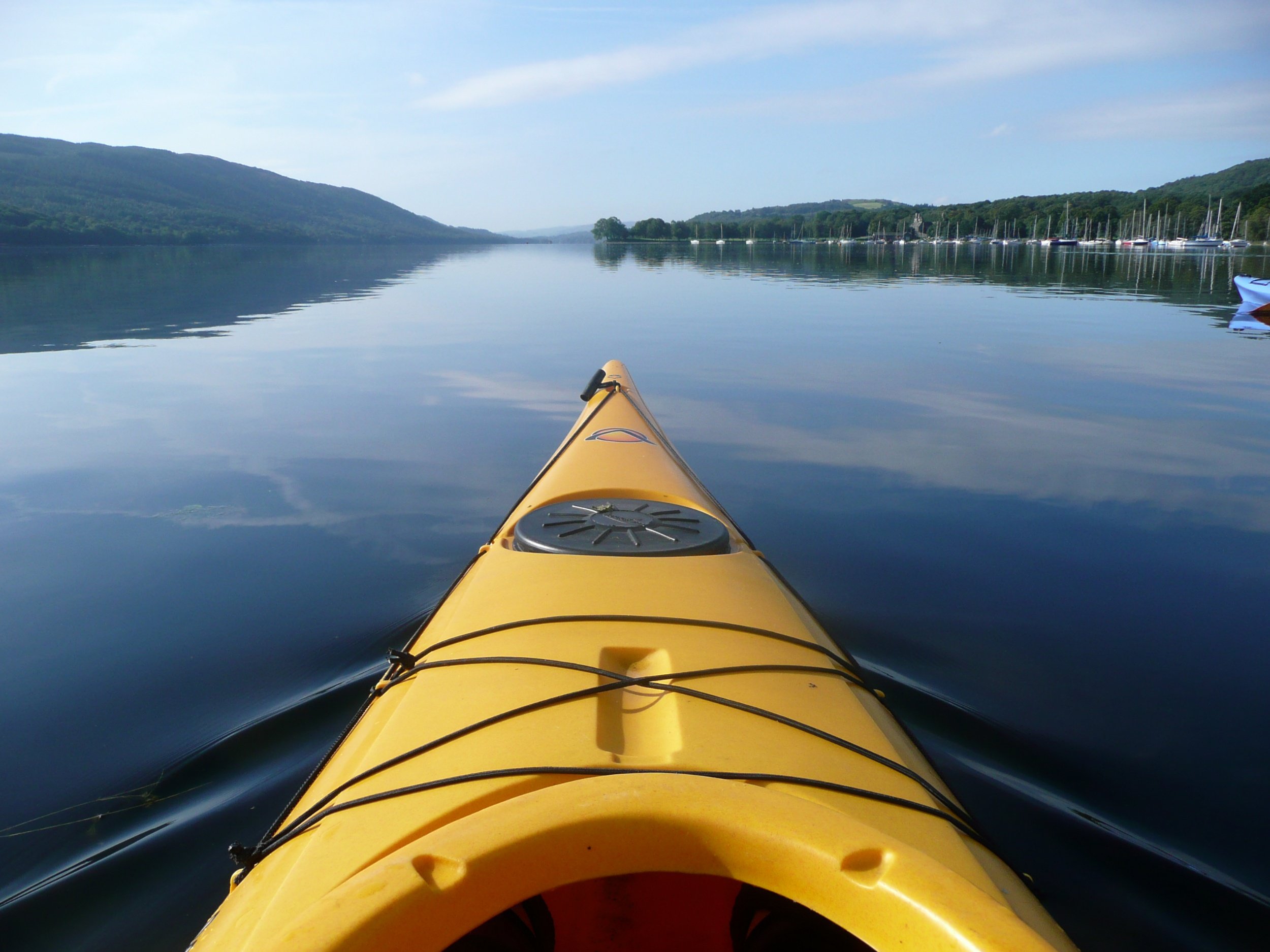 Yellow kayak on calm lake with distant hills and boats docked along the shoreline.