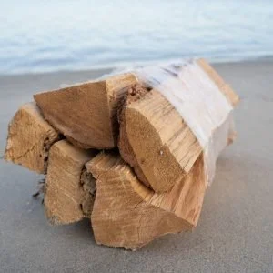 Bundle of firewood wrapped in plastic on a sandy surface near a body of water.