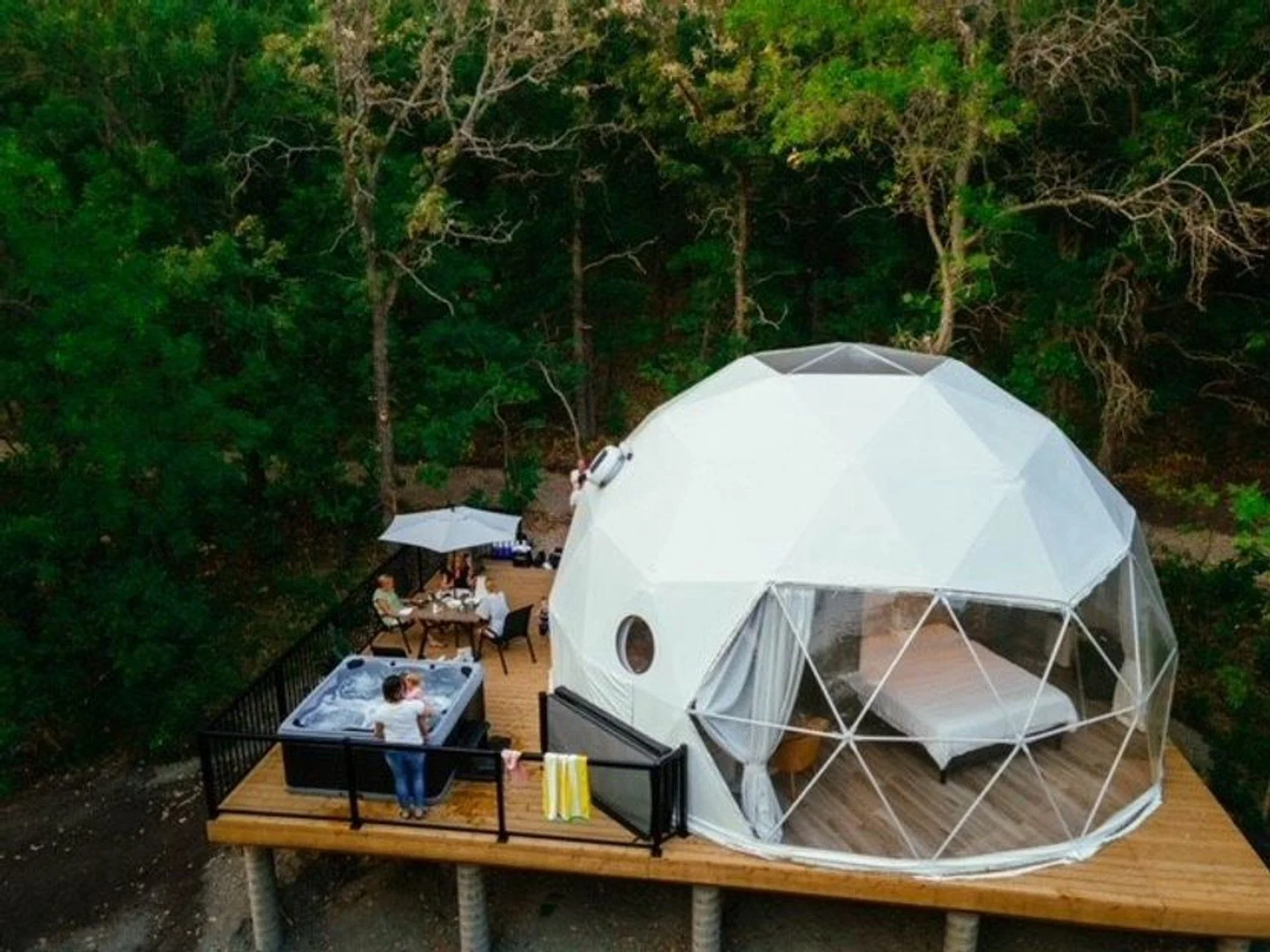 Glamping dome on elevated wooden deck with hot tub and outdoor dining area surrounded by trees.
