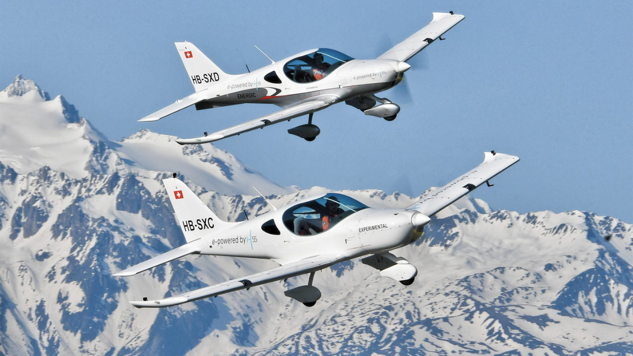 Dos aviones de entrenamiento eléctricos H55 blancos volando en formación sobre montañas suizas nevadas bajo un cielo azul.