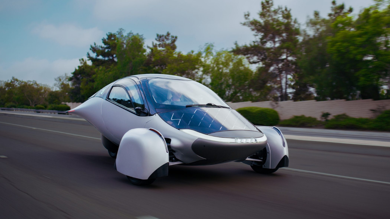 Aptera solar plateado de tres ruedas circulando por la carretera, destacando su diseño aerodinámico y futurista.