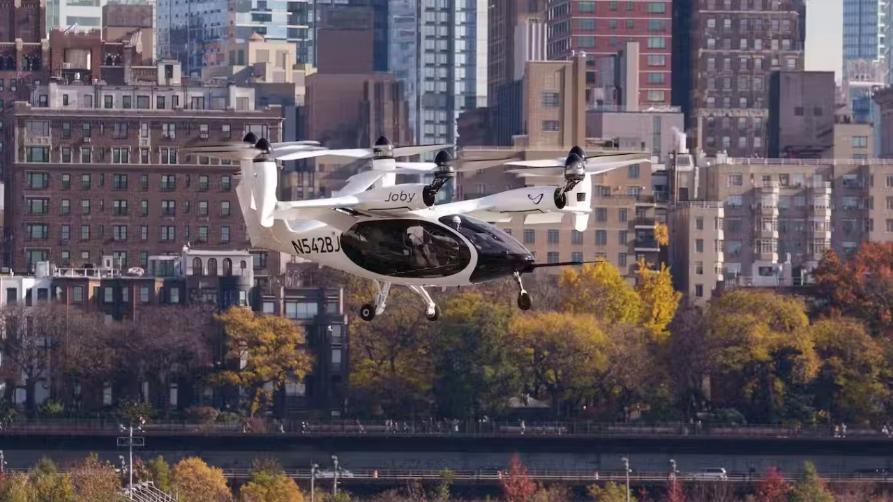 Aeronave eléctrica blanca Joby S4 volando sobre Nueva York y árboles de otoño para movilidad aérea urbana.