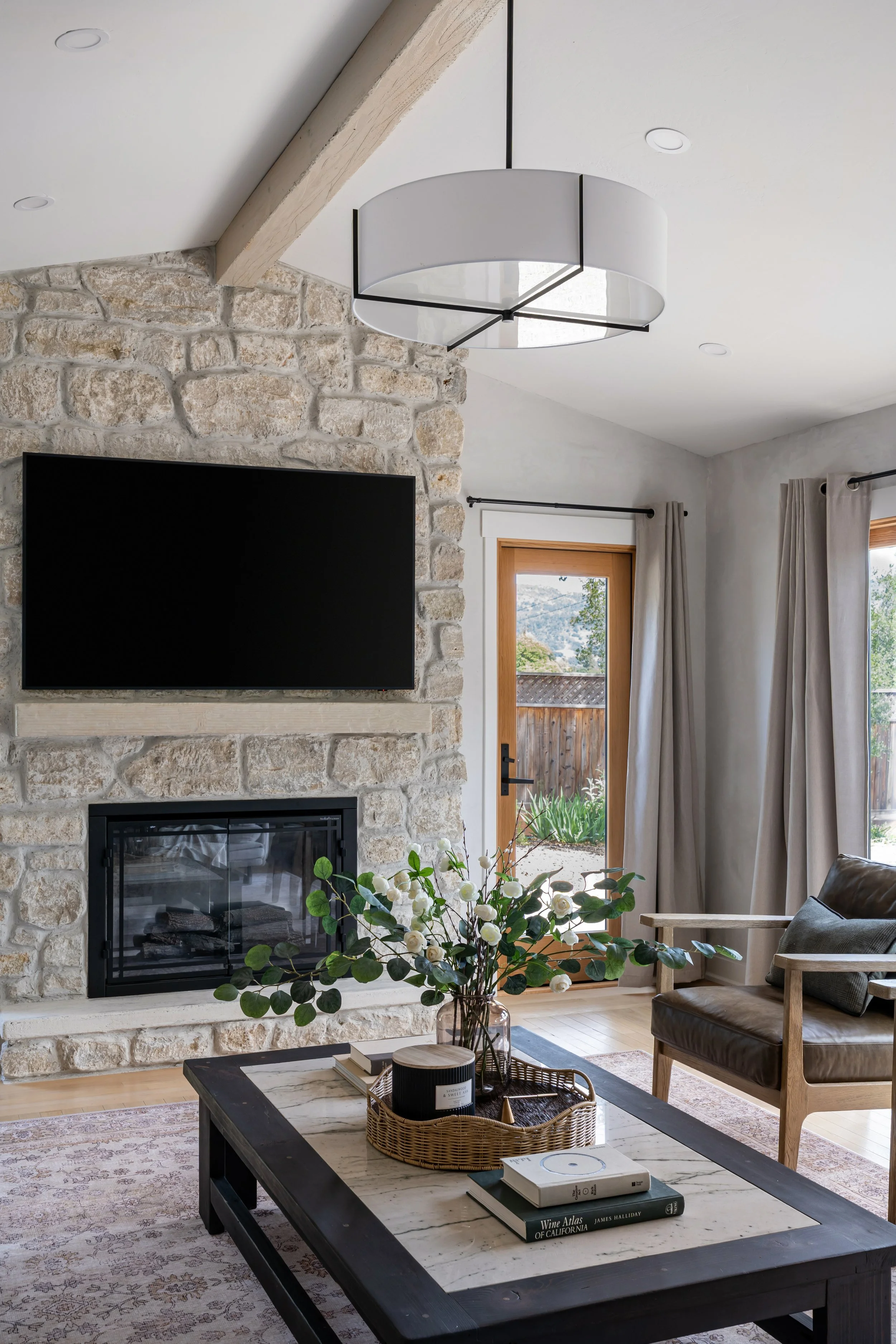 Living room with a stone fireplace, mounted flat screen TV, a coffee table with books, a vase of white flowers, a tray, and a basket. There is a armchair with a pillow, and windows with curtains showing a garden outside. A modern ceiling light hangs from the ceiling.