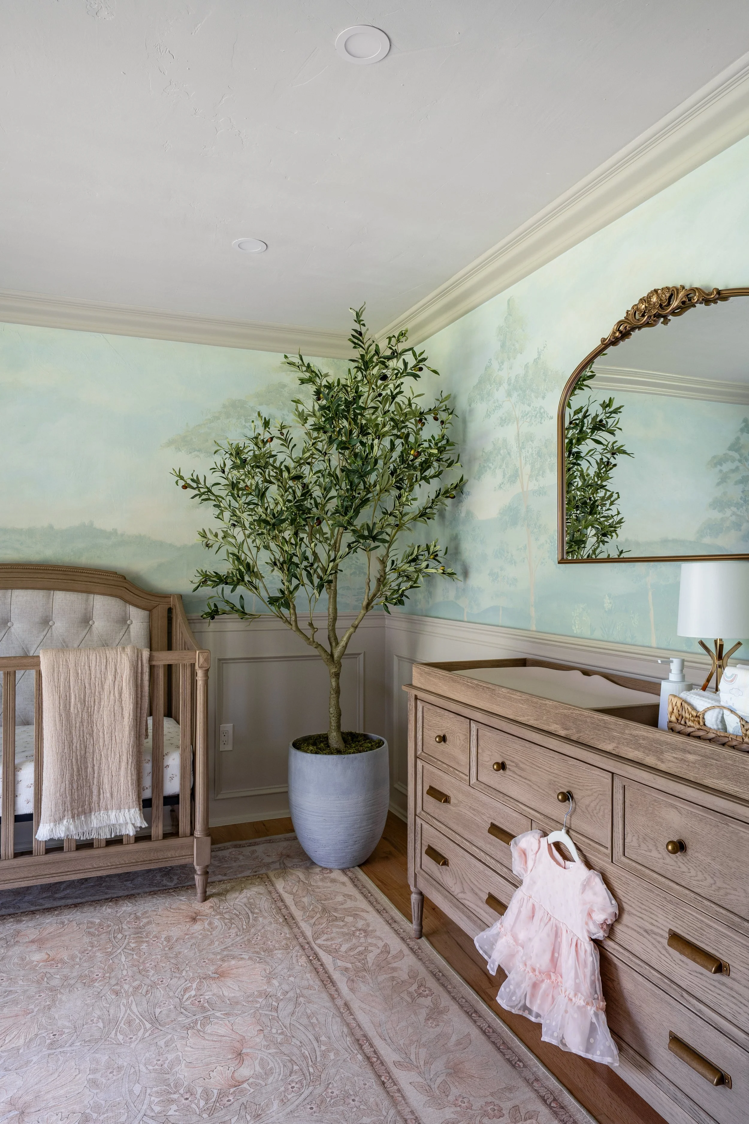 A bedroom nursery corner with a large potted tree, a wooden dresser with a dress hanging on it, a mirror with a decorative brass frame, a crib with a beige headboard, and a patterned rug. The wall has a mural of trees and a pale green background.