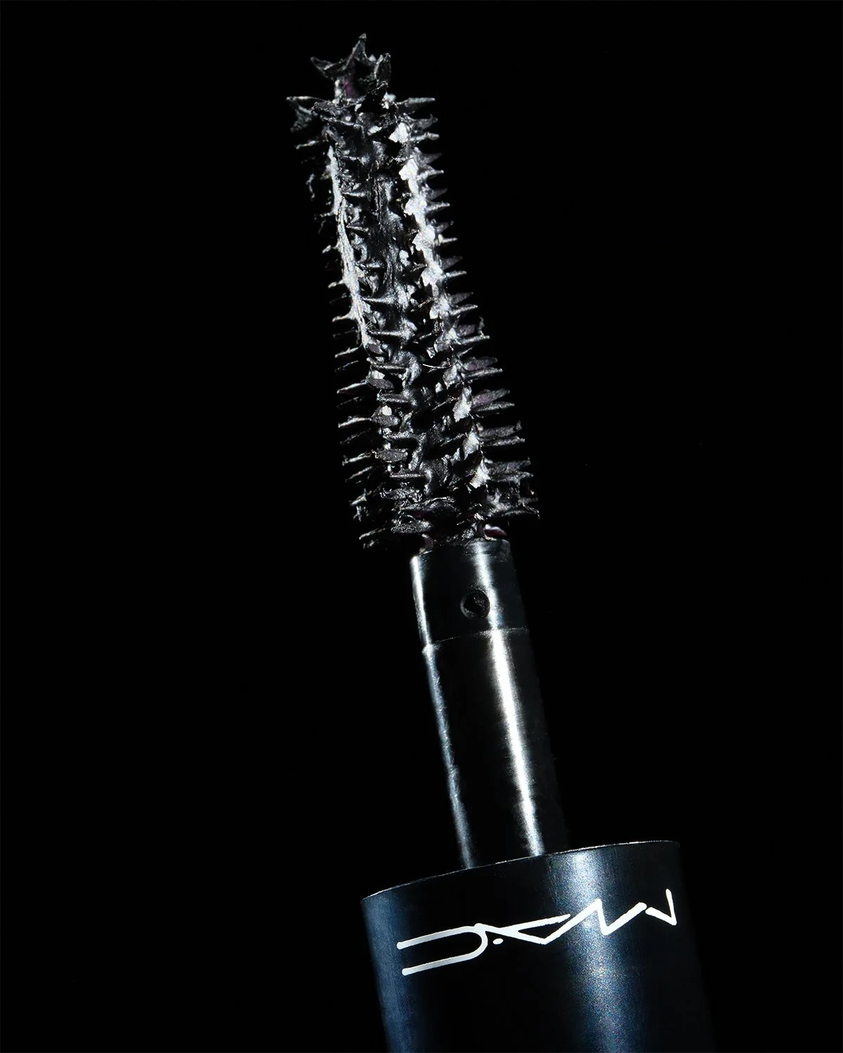Close-up of a black mascara wand with a metallic handle, displaying a textured, spiraled brush with small bristles, against a black background.