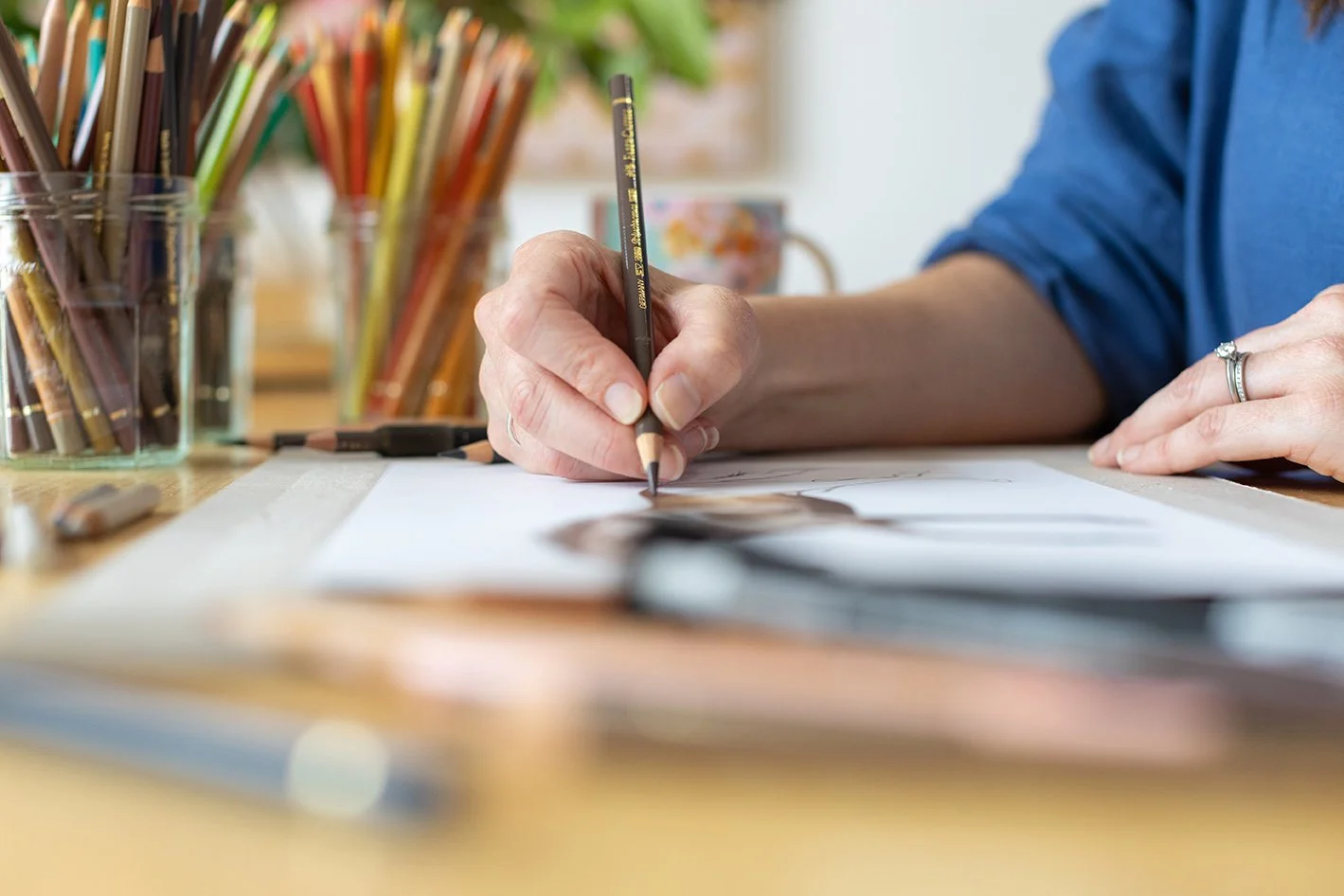 A person drawing with colored pencils on paper at a table, with containers of colored pencils in the background.