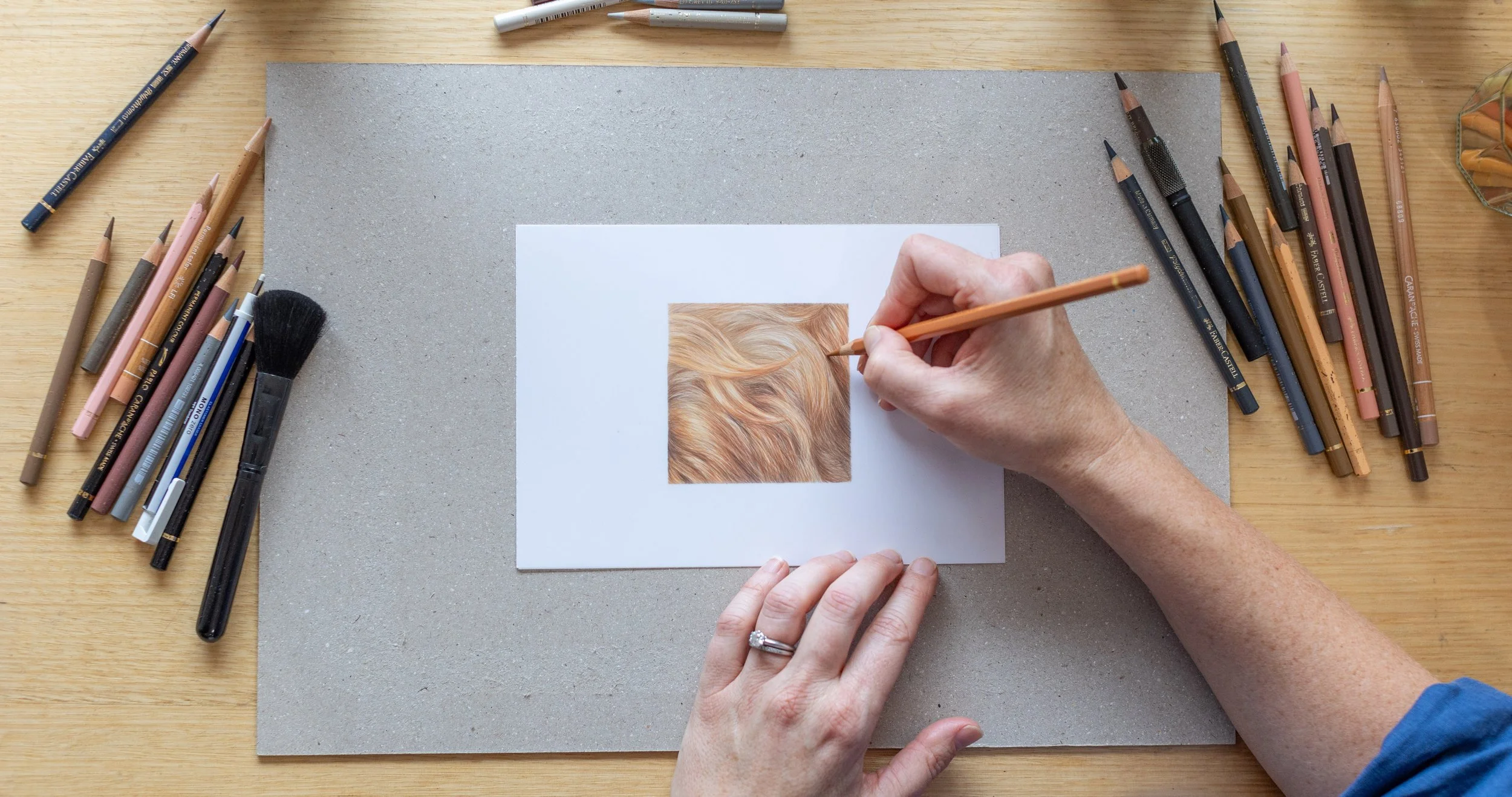 Person drawing a realistic hair texture on a small square paper using colored pencils, surrounded by various pencils and art supplies on a wooden table.